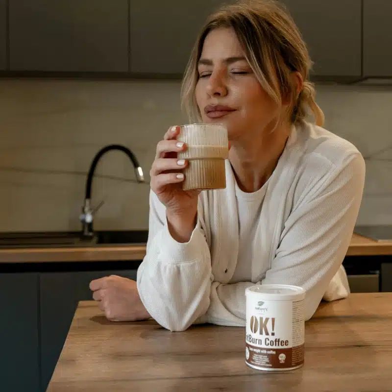 Une femme boit du café à côté d'une boîte OK! FatBurn Coffee à une table de cuisine.