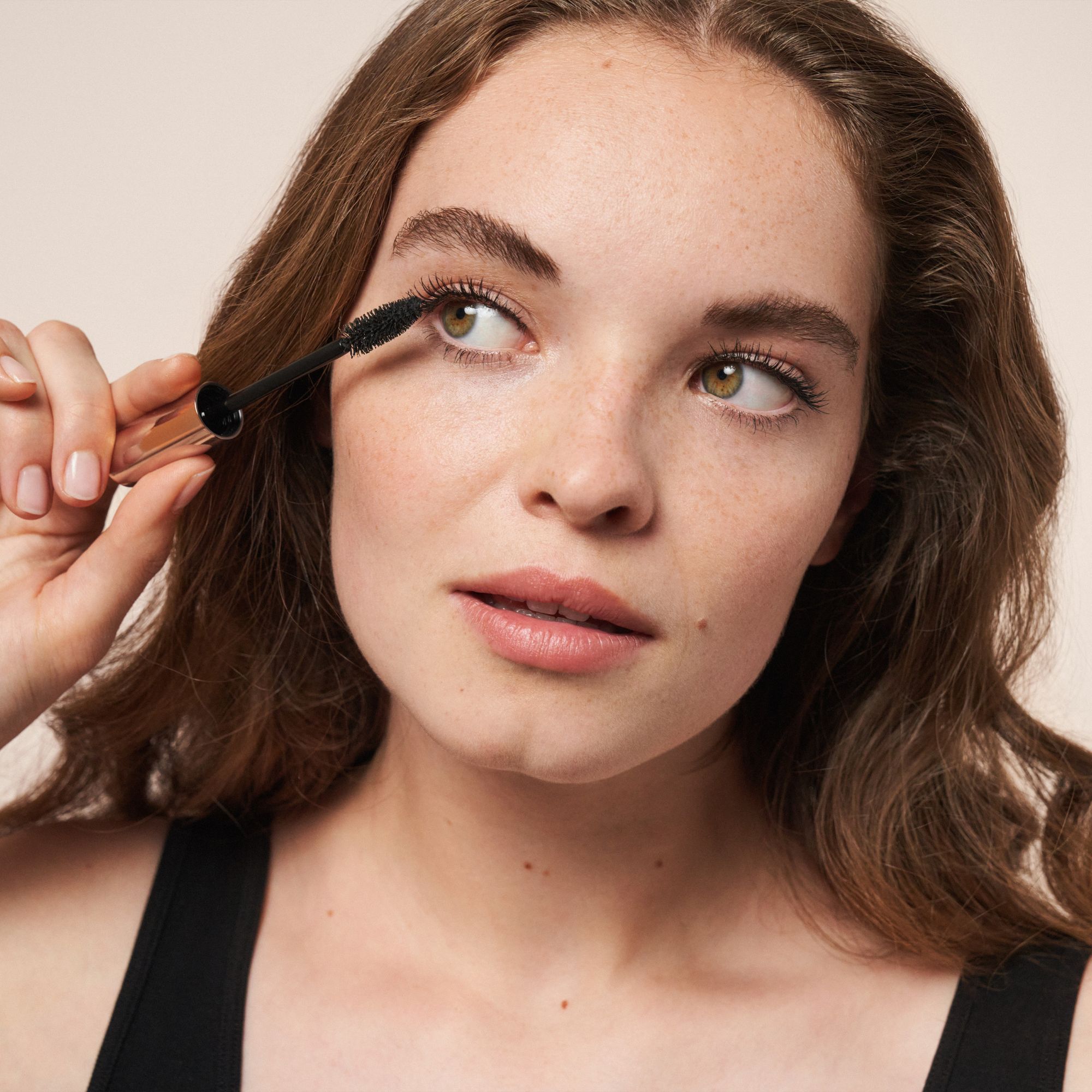 Femme appliquant du mascara. Elle tient la brosse près de l'œil. Regard vers le haut. Débardeur noir.