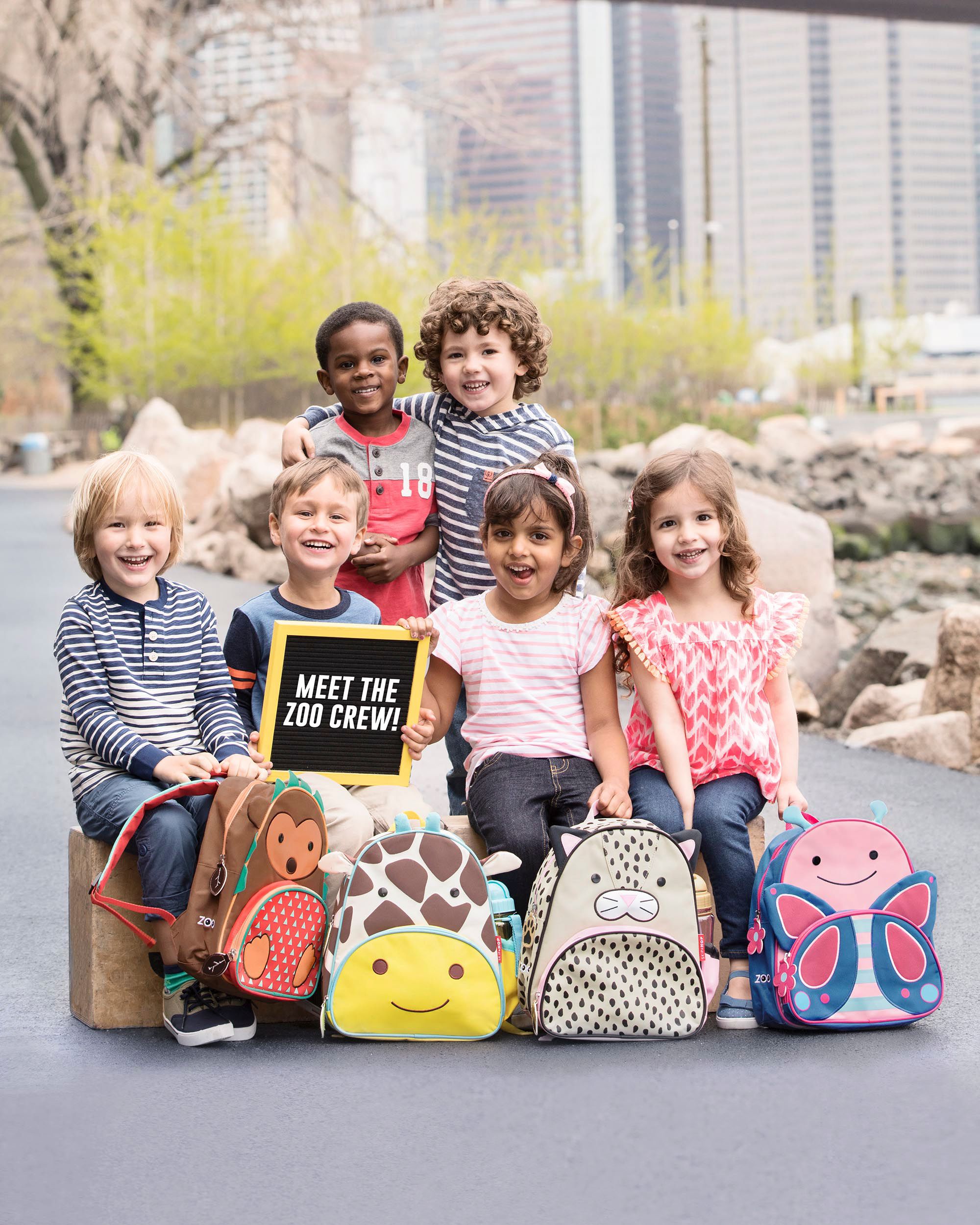 Kinderen met rugzakken. Verschillende dieren: aap, giraf, kat, vlinder. Kinderen houden een bord met "Meet the Zoo Crew!" vast.