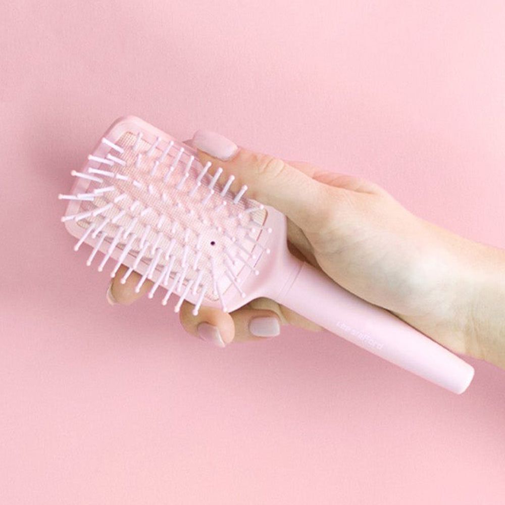Une main tient une brosse à cheveux rose pâle avec des picots blanc-rose sur fond rose.