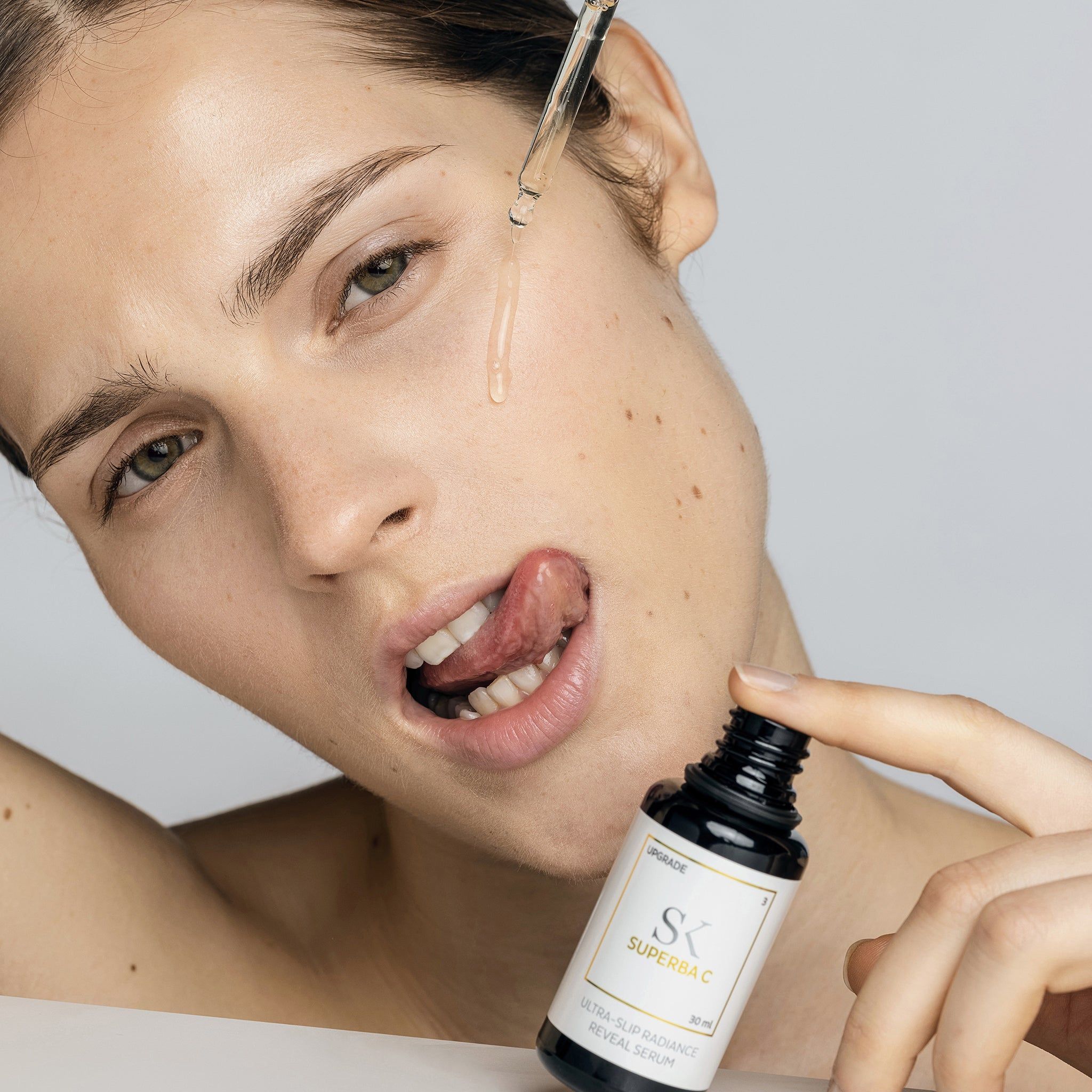 Vrouw met fles en pipet. Druppels op de tong. Op de fles staat "SK SUPERBA C" en "ULTRA-SLIP RADIANCE REVEAL SERUM".