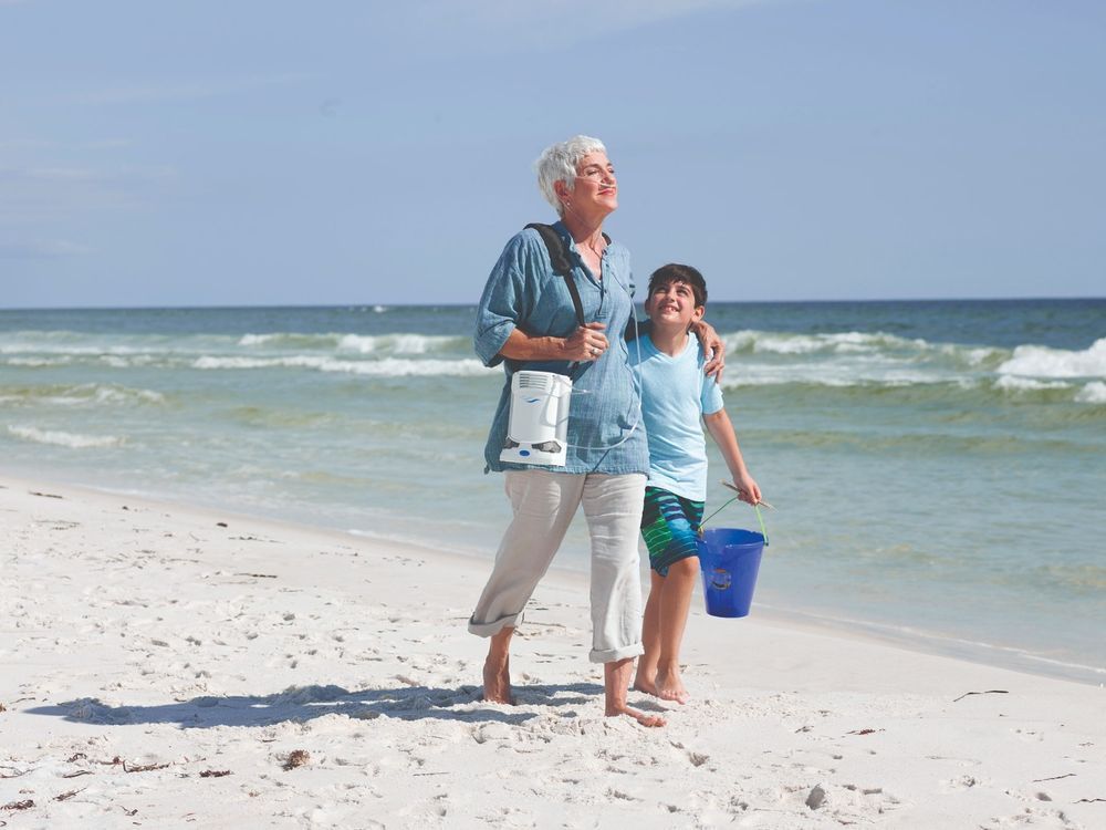 Oudere vrouw en kind op het strand, beiden met zuurstofconcentrator. Vrouw draagt apparaat. Strand en zee op de achtergrond.
