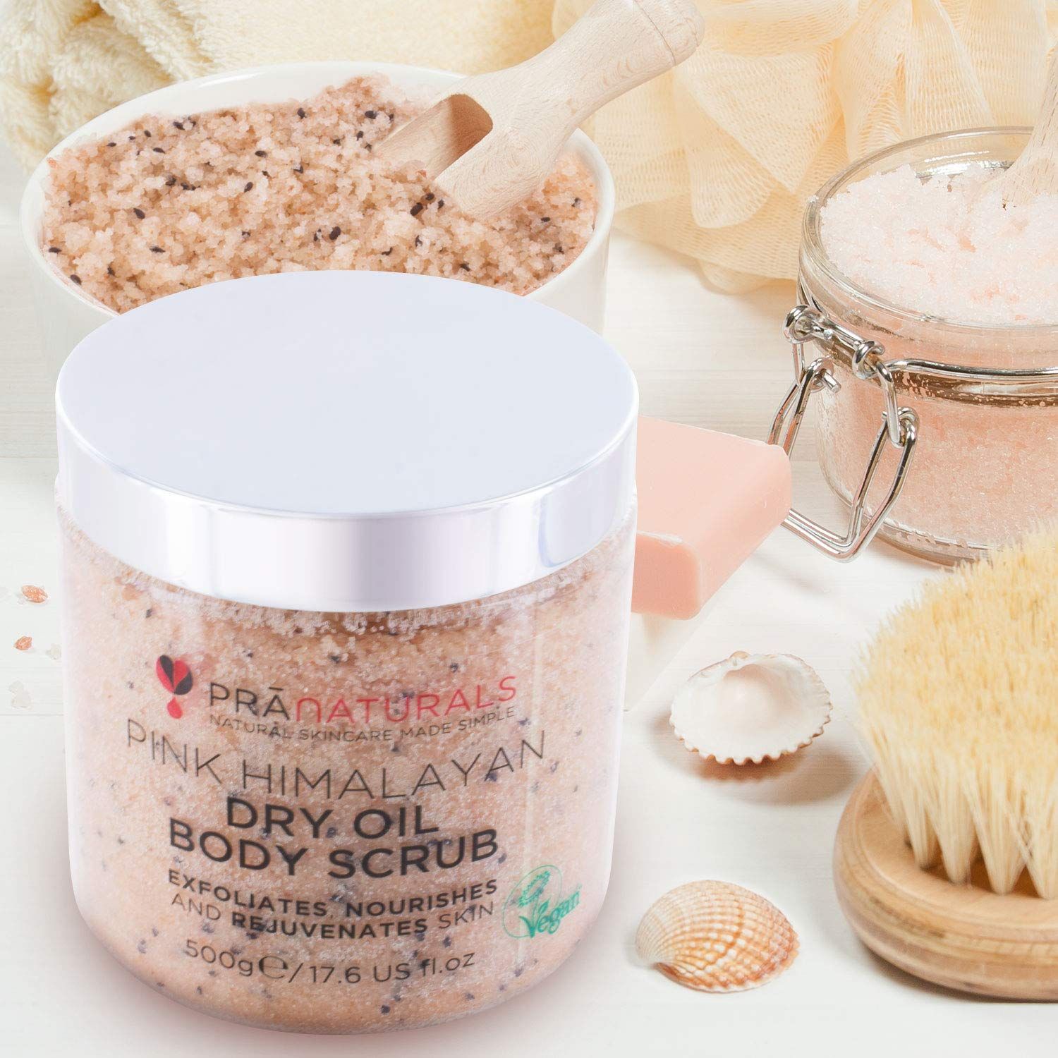 PraNaturals bodyscrub in een pot, omringd door accessoires. Tekst: Pink Himalayan Dry Oil Body Scrub. Op witte ondergrond.