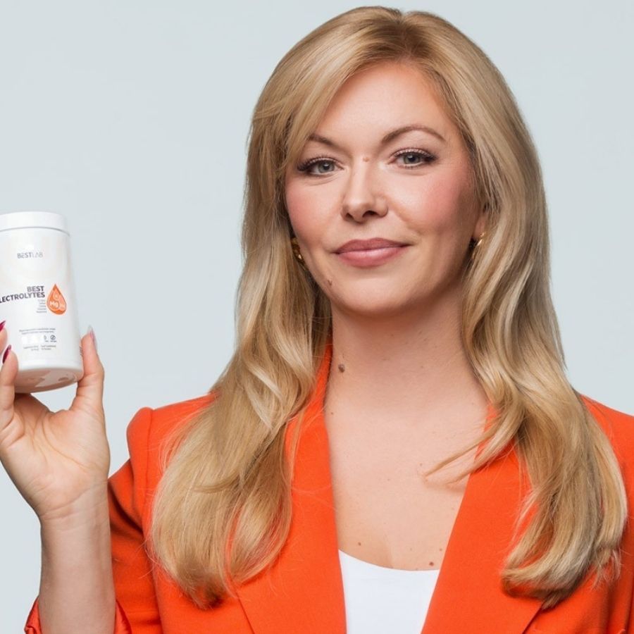 Femme tenant un conteneur blanc BEST ELECTROLYTES. Elle porte un blazer orange. Cheveux blonds.