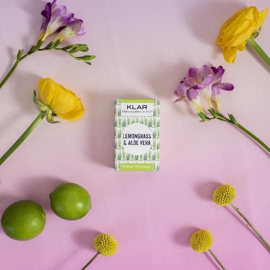 Klar-Seifen verpakking, omringd door bloemen en limoenen. Verpakking: groen-wit, logo Lemongras & Aloe Vera. Tekst: Vast Shampoo. Merk: Klar.