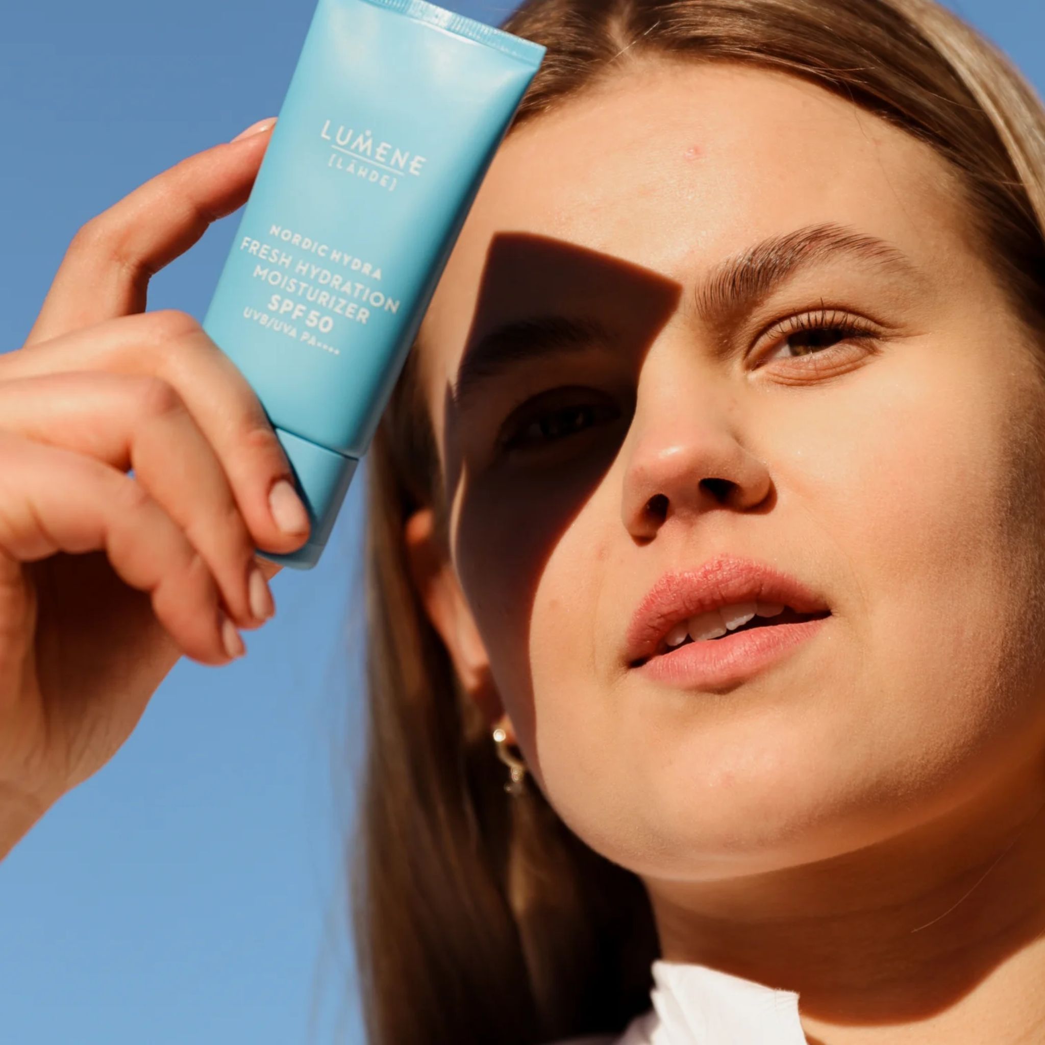 Femme tenant un tube bleu clair devant un ciel bleu. Texte: LUMENE [LÄHDE], Nordic Hydra Fresh Hydration Moisturizer SPF50. Visage à l'ombre.