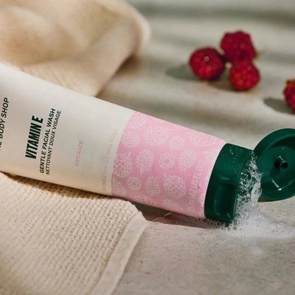 The Body Shop Vitamin E Gentle Facial Wash, tube ouvert, mousse sortant. Framboises et serviette en arrière-plan.