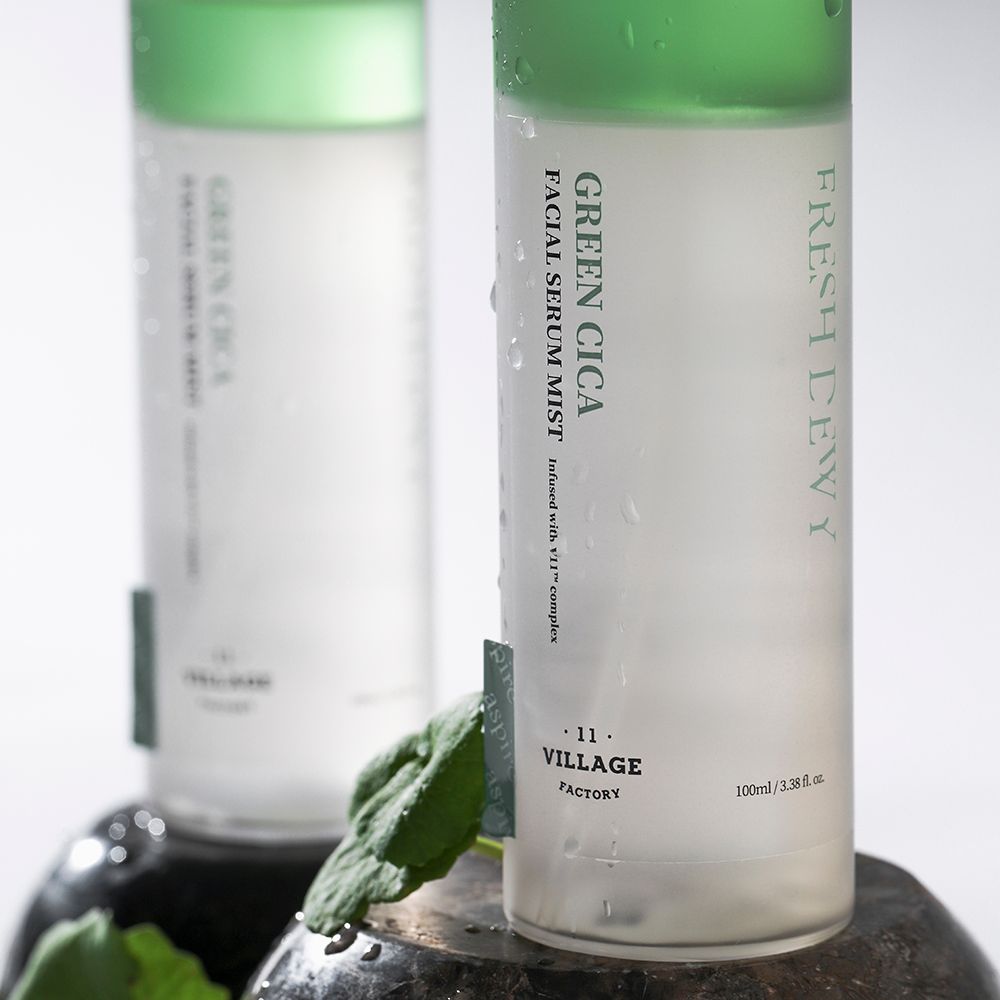 Twee flessen met groene band en witte dop. Opschrift: Green Cica Facial Serum Mist. Merk: Village 11 Factory. Geplaatst op stenen.