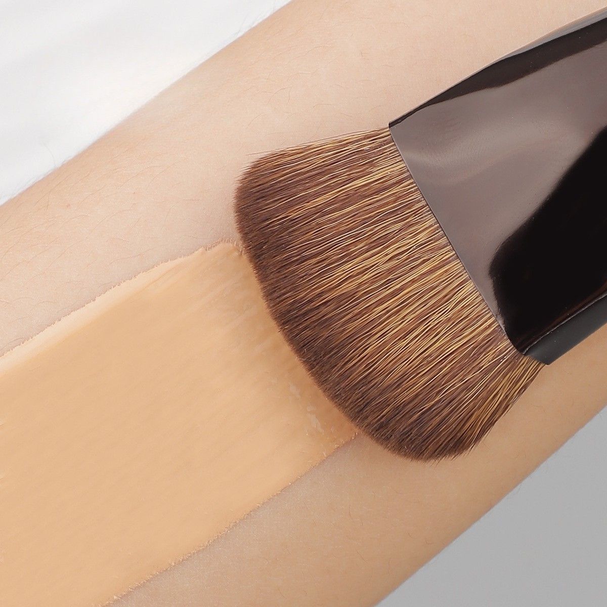 Un pinceau de maquillage plat aux poils brun clair applique un fond de teint liquide beige clair sur une peau claire.