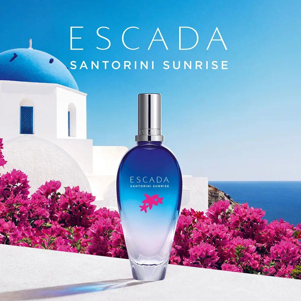 Blauw-transparante fles voor Griekse achtergrond. Opschrift: ESCADA SANTORINI SUNRISE. Roze bloemen.
