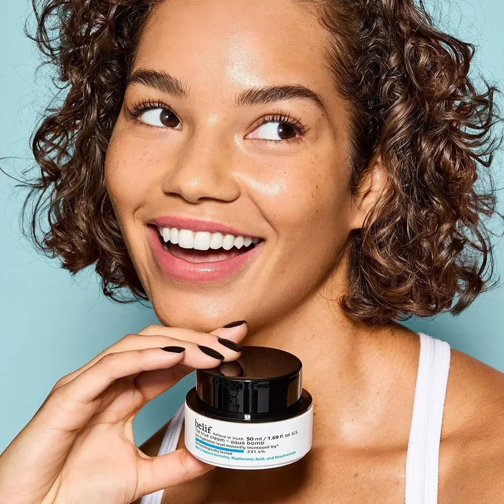 Femme souriante aux cheveux bouclés tenant un pot de Belif The True Cream Aqua Bomb.