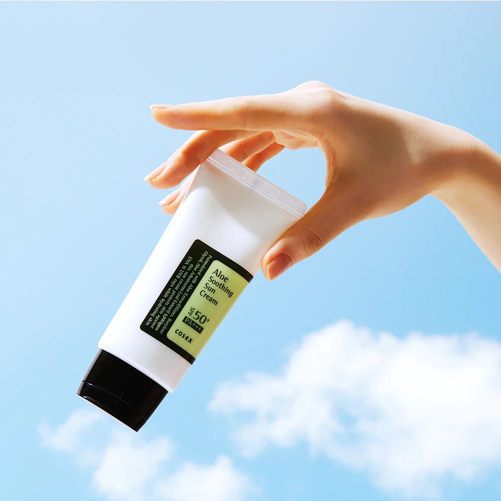 Witte tube met zwarte dop in hand, blauwe lucht. Opschrift: Aloe Soothing Sun Cream, COSRX.