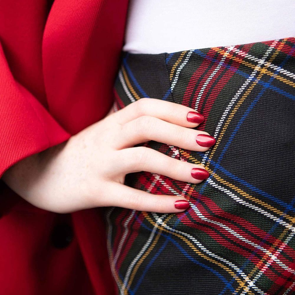Hand met gelakte nagels. De nagels zijn rood en glanzend. Persoon draagt rode jas en geruite rok.