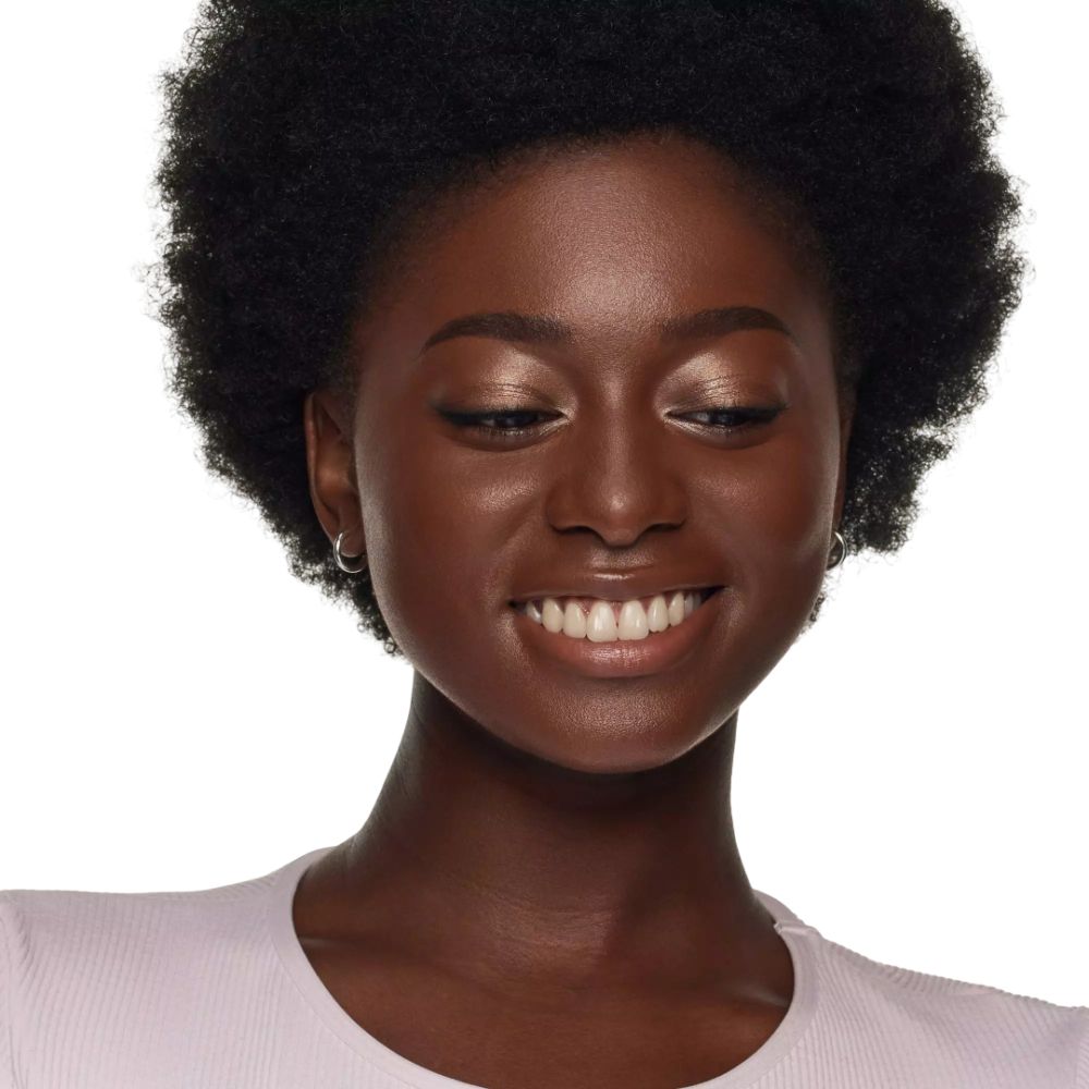 Portrait d'une femme à la peau foncée souriante. Elle porte de l'ombre à paupières claire.