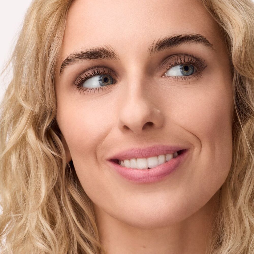 Portret van een vrouw met blond krullend haar, die opzij kijkt en glimlacht. Roze lippenstift.