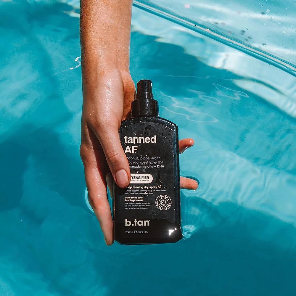 Zwarte sprayfles 'tanned AF' in een hand boven blauw water. 'b.tan' is zichtbaar. Bevat kokos, jojoba, argan, avocado, rozenbottel, druif & macadamia oliën + DHA.