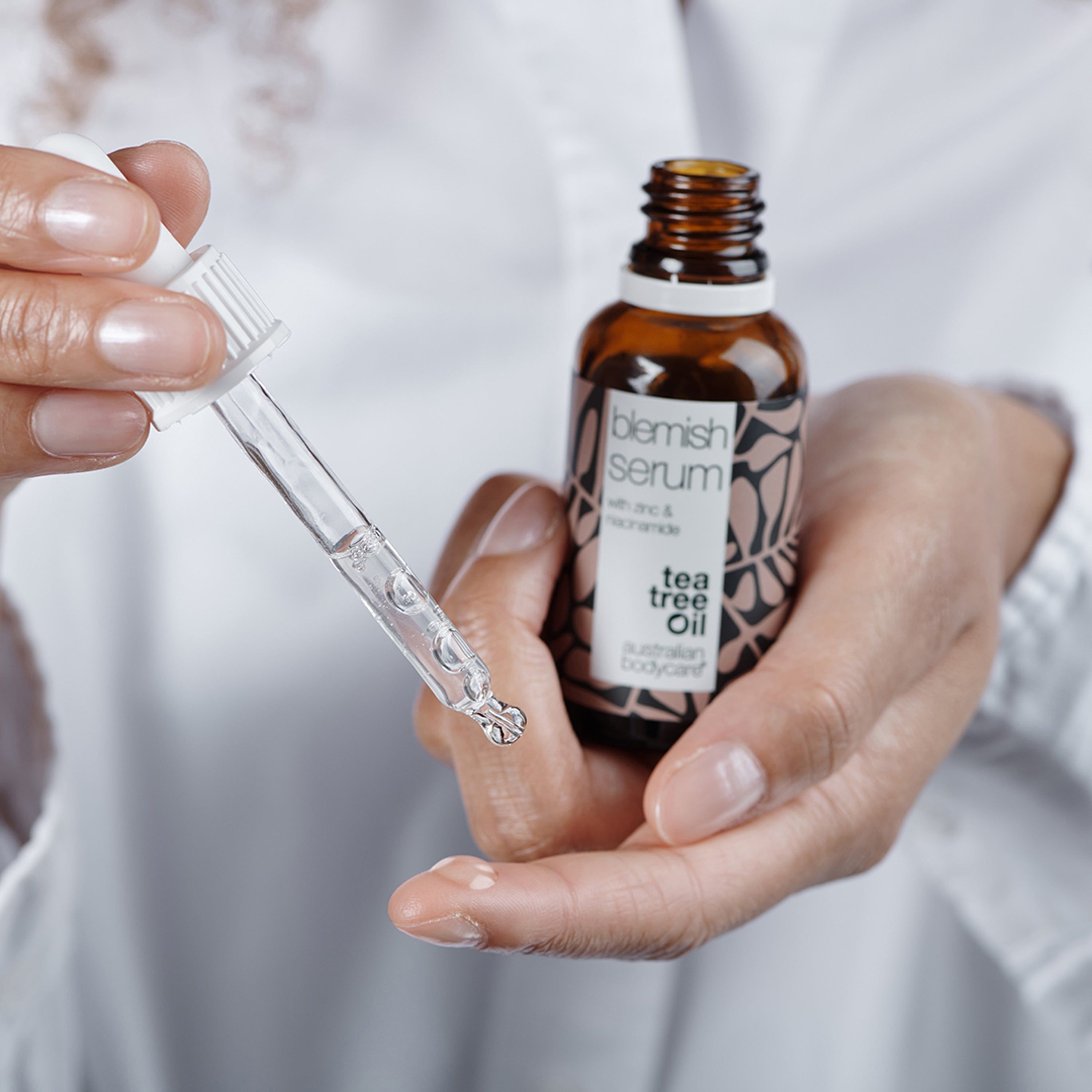 Handen houden serumfles en pipet. Opschrift: Blemish Serum, Tea Tree Oil, Australian Bodycare.