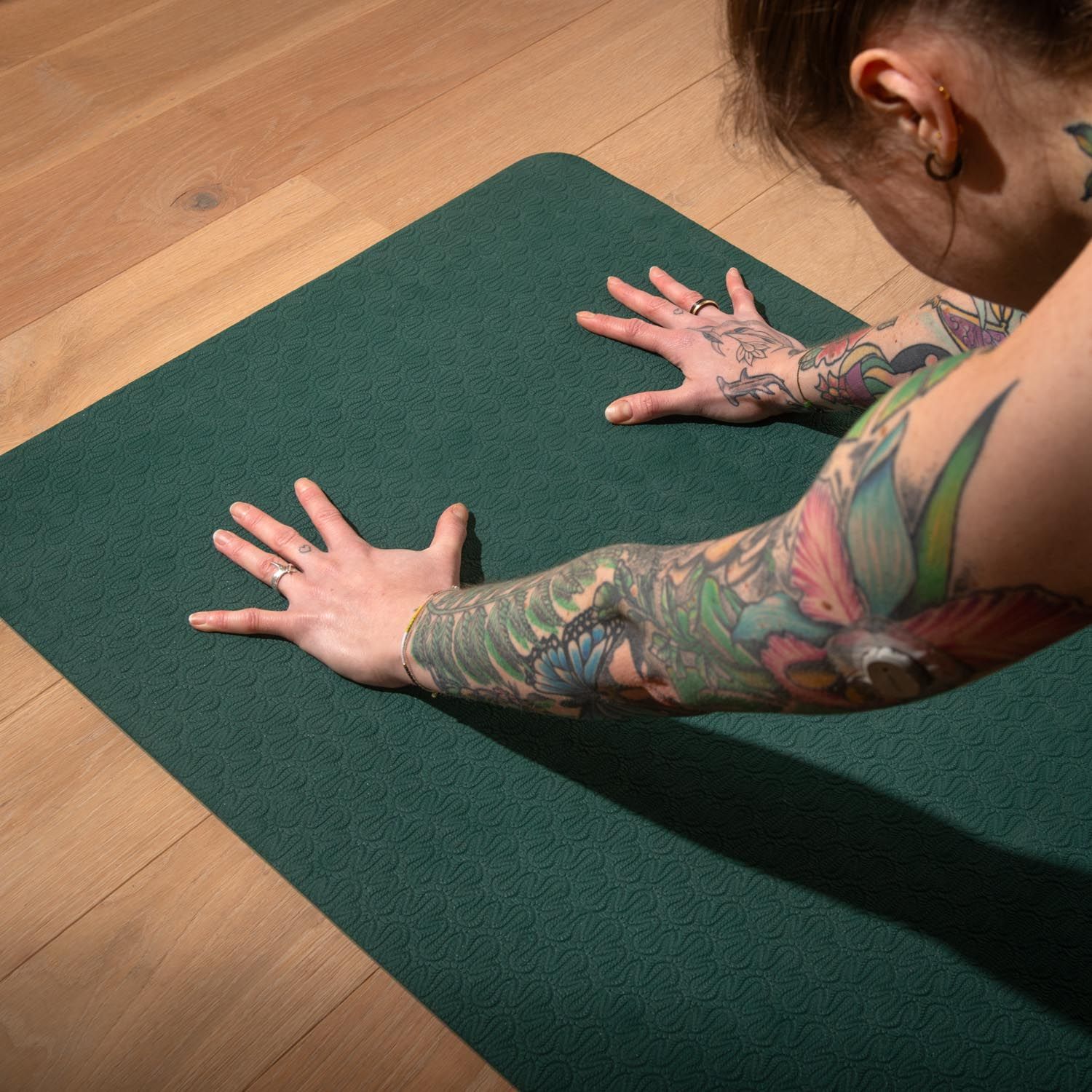 Persoon ligt op een donkergroene yogamat. Armen en handen uitgestrekt. Houten vloer op de achtergrond.