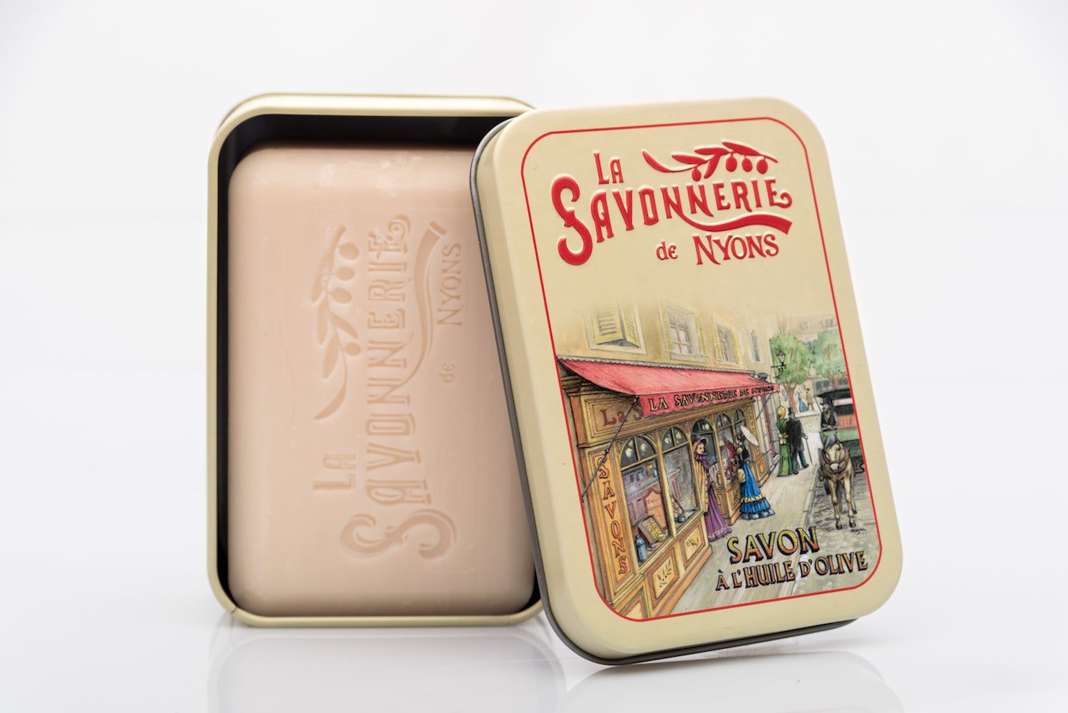 Rechthoekige metalen doos met zeep. Doos open, zeep zichtbaar. Opschrift: La Savonnerie de Nyons. Illustratie van een boetiek.