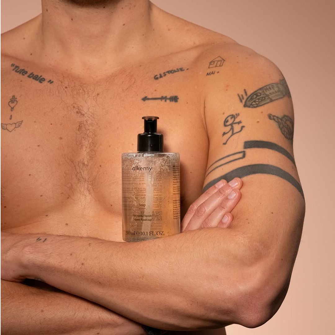 Man met tatoeages houdt fles heldere vloeibare zeep voor de borst.