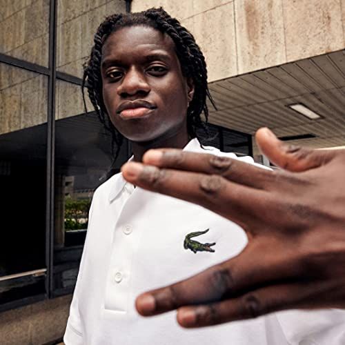 Un jeune homme avec des tresses porte un polo blanc avec un logo crocodile vert. Un bâtiment en arrière-plan.