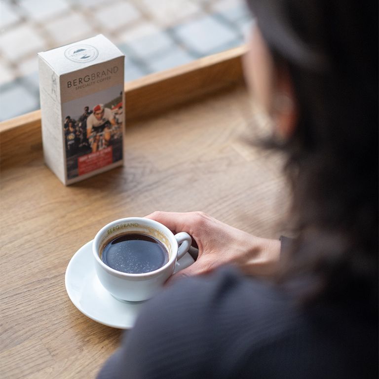 Koffiekopje met koffie en koffieverpakking BERGBRAND Eddy would Attack. Persoon op de achtergrond. Op een tafel.