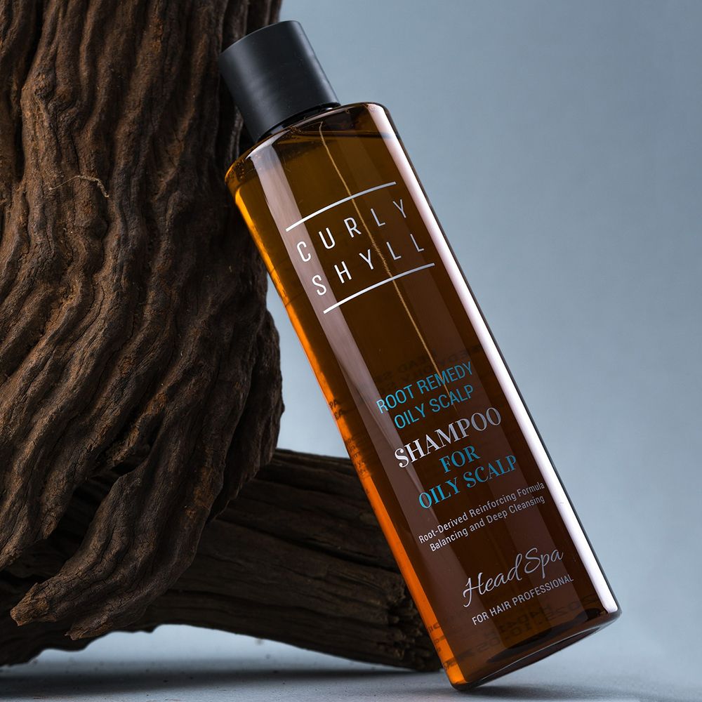 Flacon de shampooing brun, placé en biais devant du bois. Inscription : CURLY SHYLL, Root Remedy Oily Scalp Shampoo. Texte : For Oily Scalp, Head Spa.