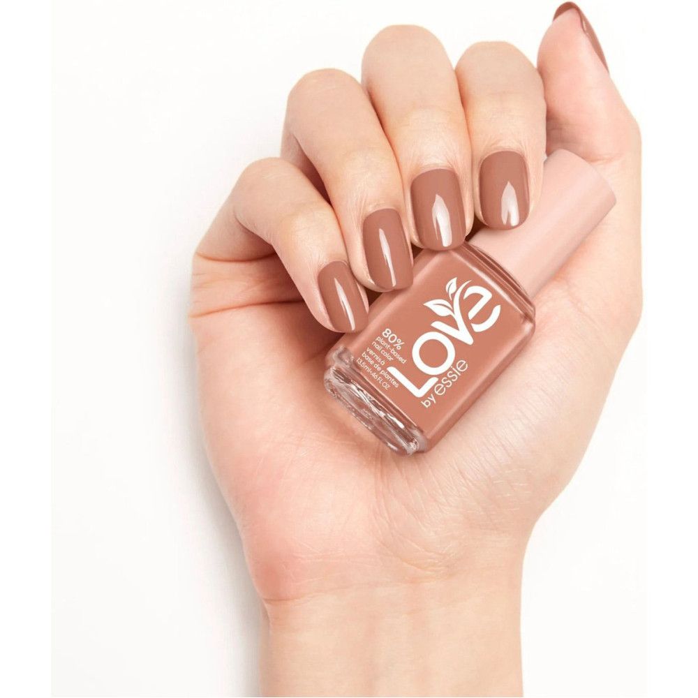 Main tenant un flacon de vernis à ongles. Ongles vernis. Inscription : Love by Essie. Bouchon rose, forme carrée.