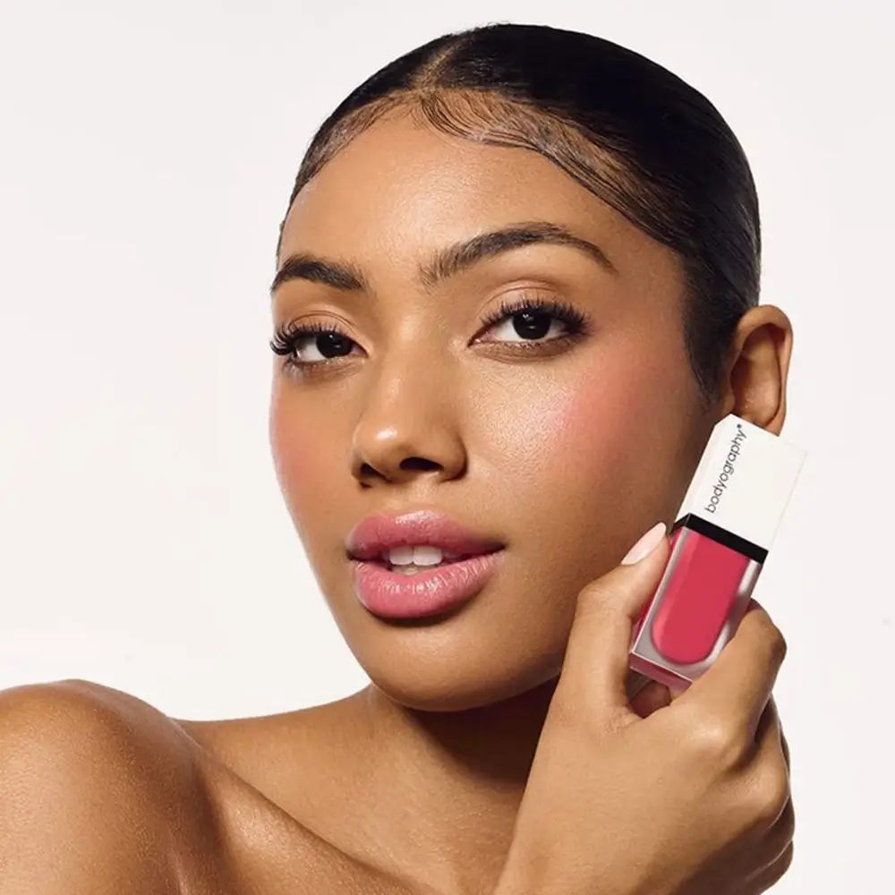 Vrouw houdt vloeibare blush vast. Product BODYOGRAPHY. Koraalroze tint. Aangebracht op wangen en lippen.