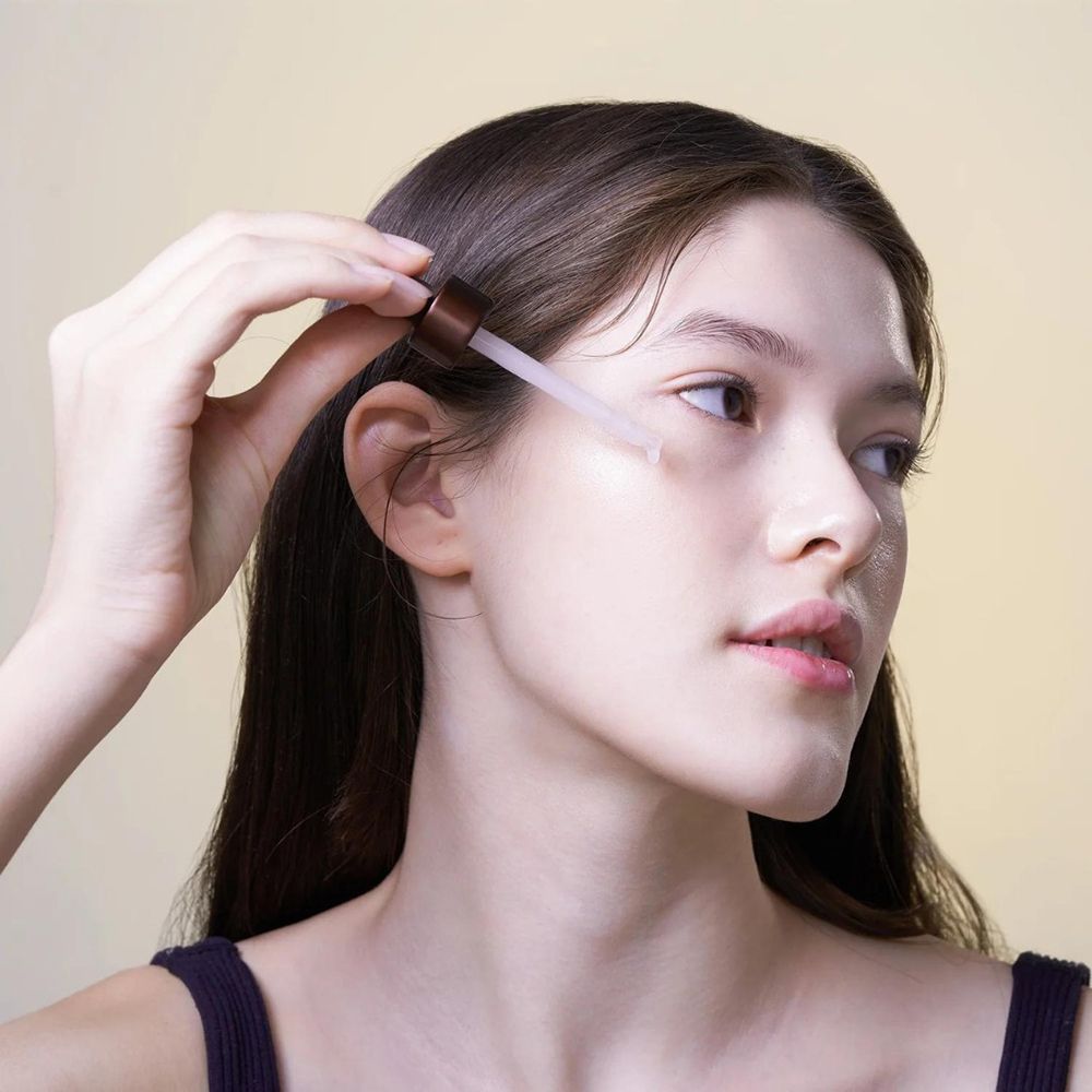Jeune femme aux longs cheveux appliquant du sérum sur son visage avec une pipette. Peau claire, regard de côté.