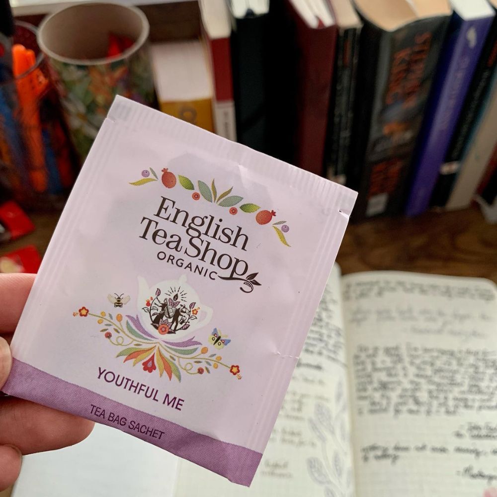 Enkele theezakje ETS - Youthful Me. Opschrift: English Tea Shop Organic. Zakje wordt vastgehouden. Achtergrond: notitieboek, boeken.