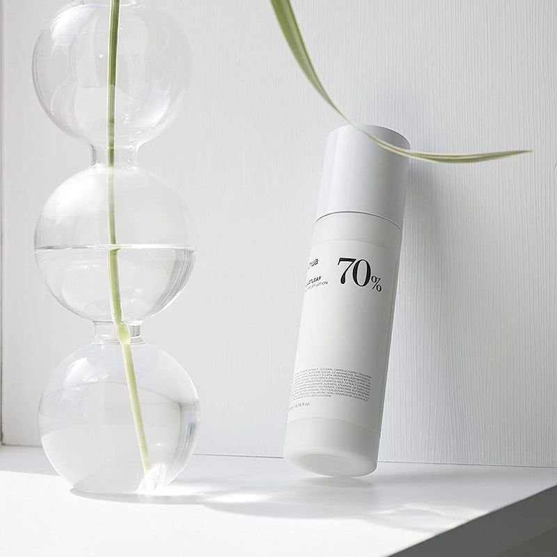 Witte lotionfles naast glazen vazen en plant. Opschrift: Anua, Heartleaf 70% Daily Releaf Lotion, 70%.