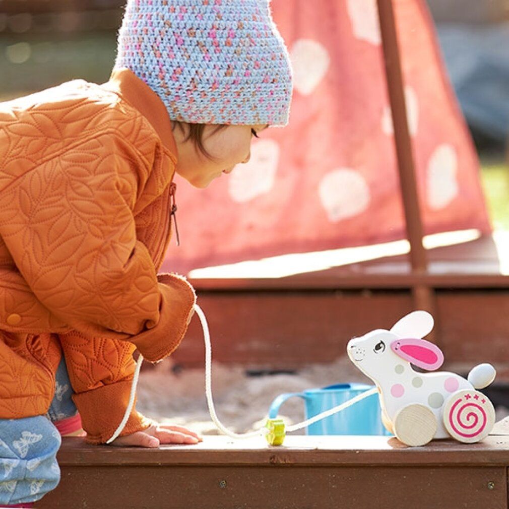 Enfant jouant avec un lapin en bois à tirer. Blanc avec pois roses et gris. Trèfle sur la ficelle. L'enfant porte un bonnet et une veste.