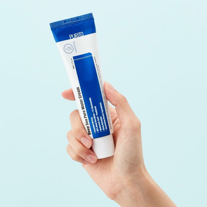 Main tenant un tube de crème. Tube bleu et blanc. Texte: Deep Sea Pure Water Cream. Marque PURITO. Fond bleu clair.