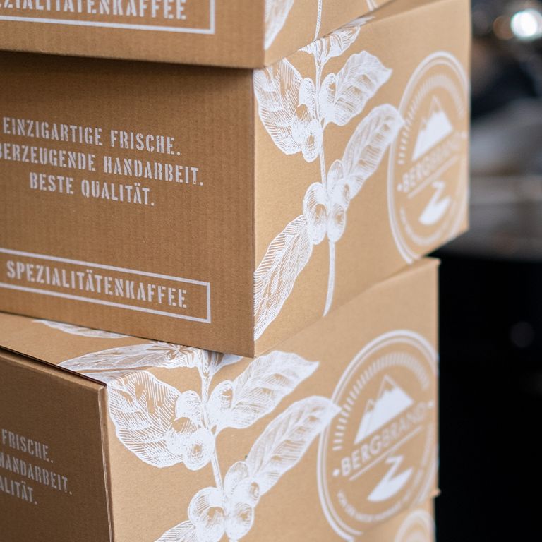 Koffieproducten gestapeld in dozen. Opschrift: BERGBRAND, Spezialitätenkaffee. Tekst: Unieke versheid, overtuigend handwerk, beste kwaliteit.