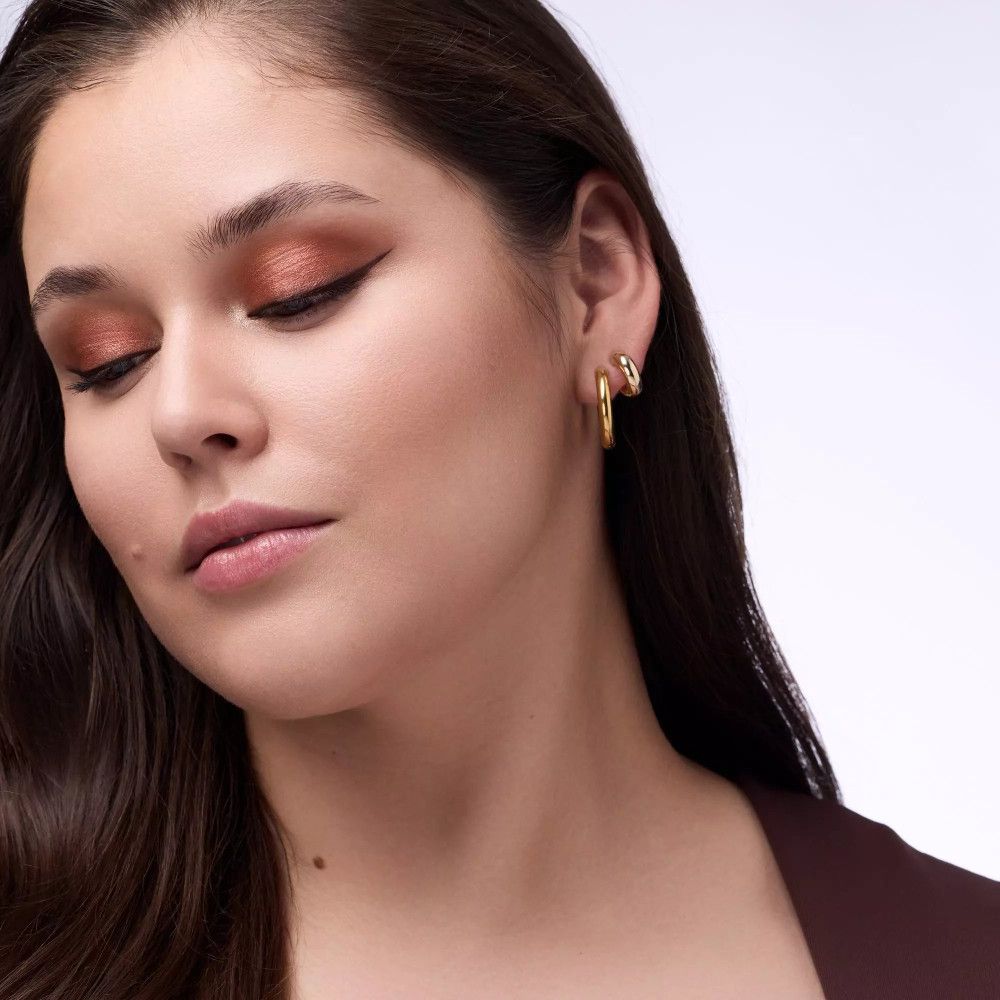 Portrait d'une femme avec fard à paupières dans les tons marron et orange. Elle porte des boucles d'oreilles dorées et regarde vers le bas.