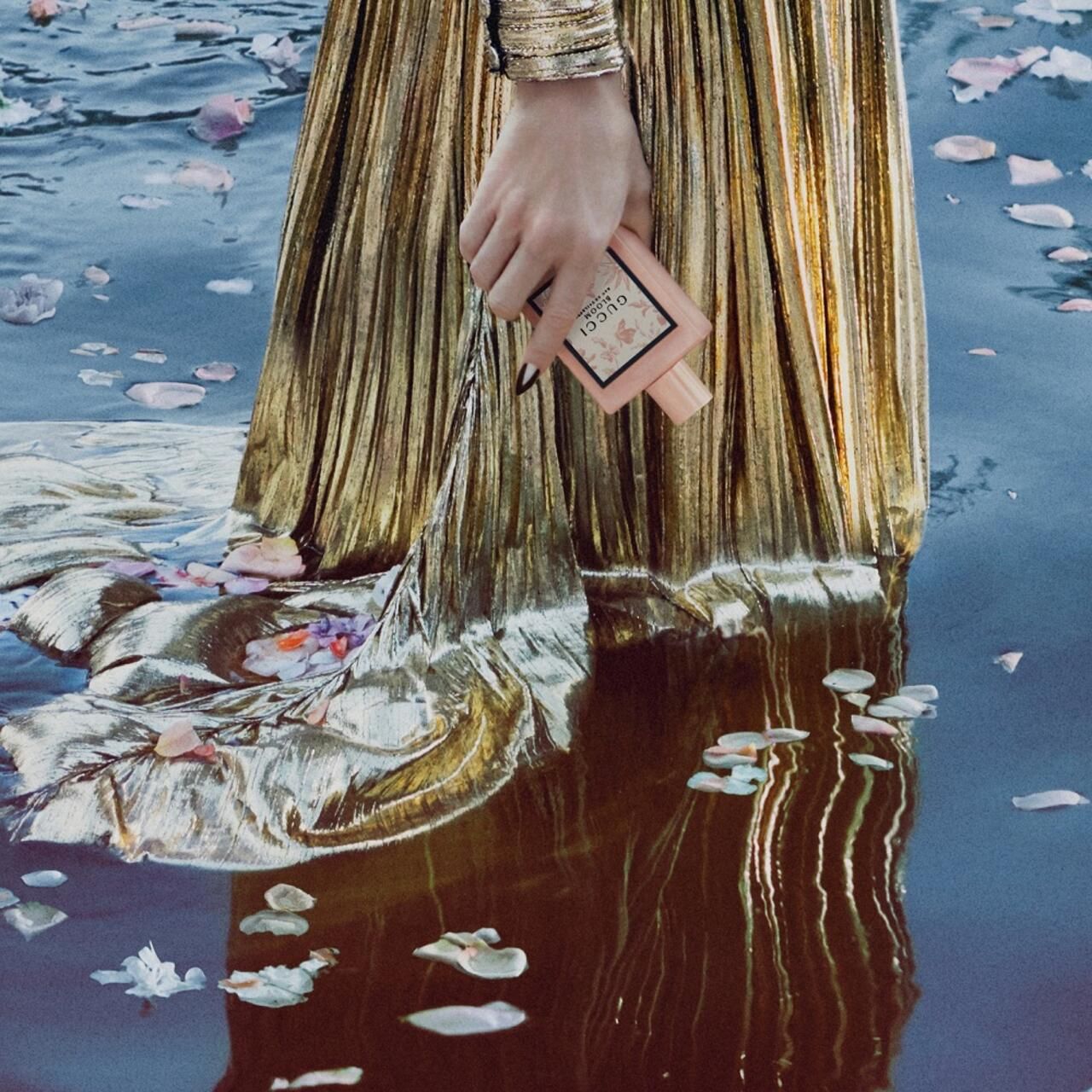 Hand houdt roze fles met bloemenprint in water. Opschrift: Gucci Bloom Eau de Toilette.
