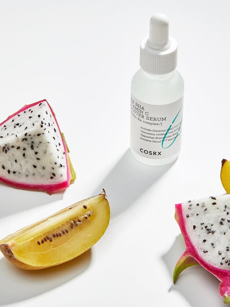Flacon de sérum avec pipette, à côté de kiwi et fruit du dragon. Inscription : AHA BHA Vitamin C Booster Serum. Marque : COSRX.