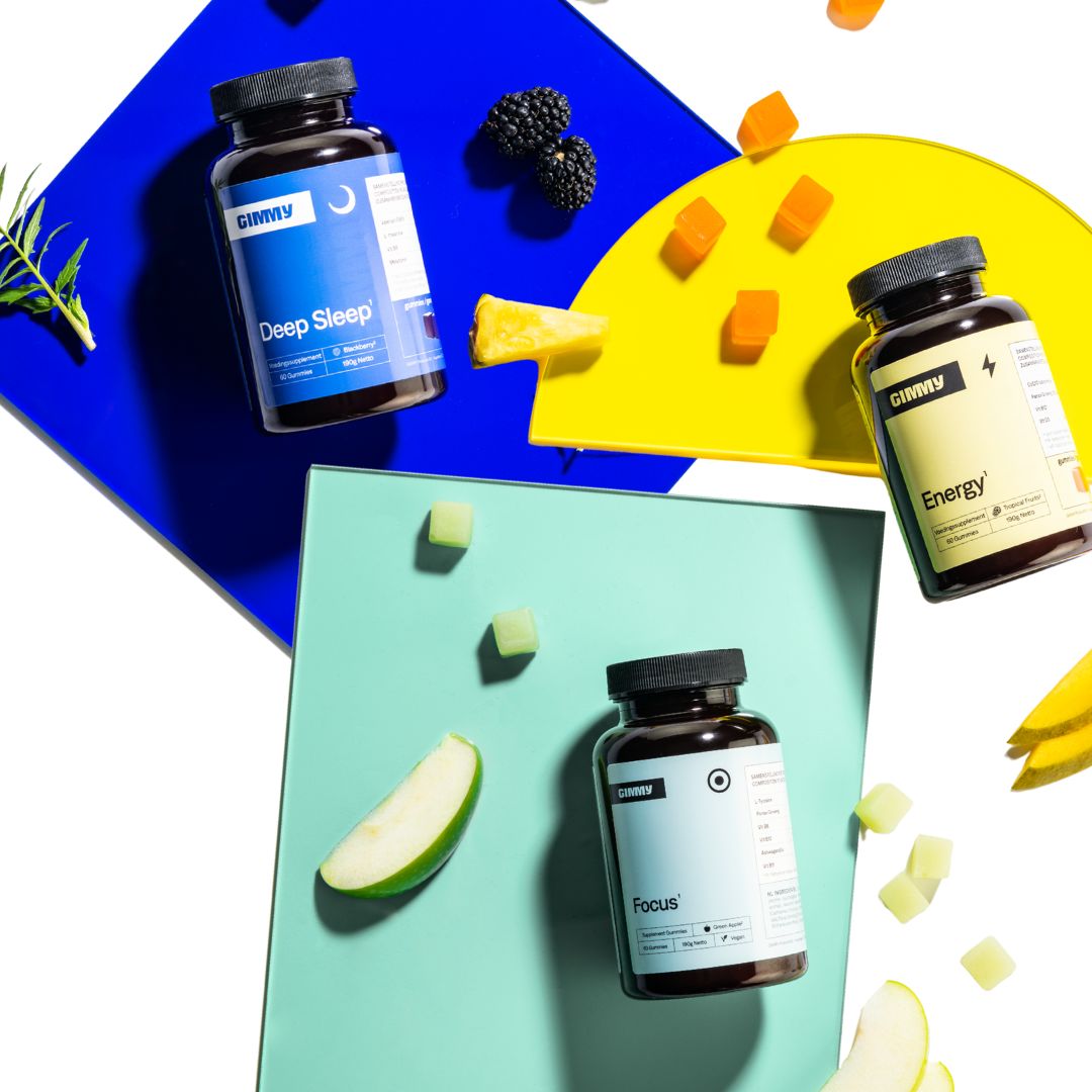 Trois flacons de compléments alimentaires GIMMY sur fond coloré. Des gommes et des morceaux de fruits sont à côté.