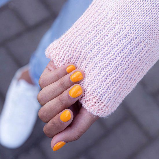 Main avec ongles vernis en orange. Pull rose clair et chaussures blanches en arrière-plan.