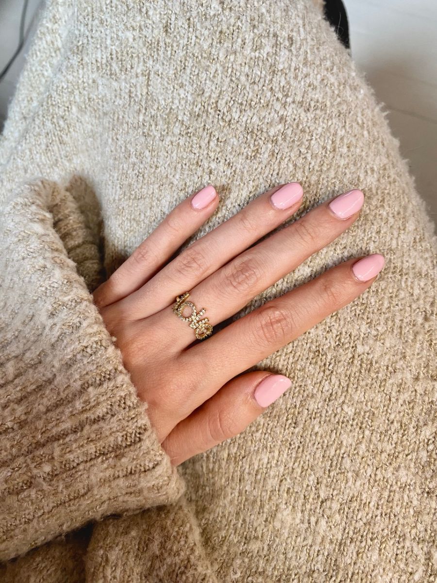 Gros plan d'une main avec des ongles peints en rose clair. La main porte une bague dorée. La personne porte un pull beige.