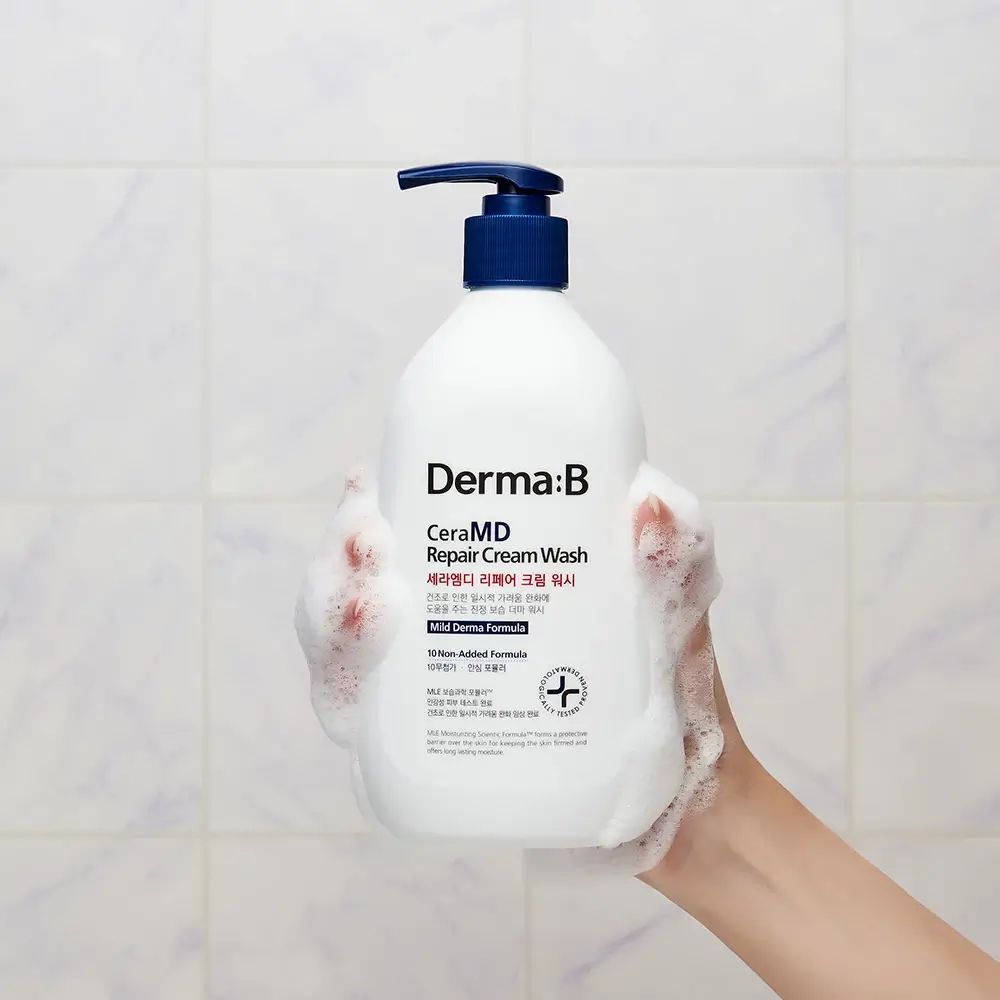 Witte fles met blauwe pomp in hand. Schuim. Opschrift: Derma:B CeraMD Repair Cream Wash. Achtergrond: tegels.
