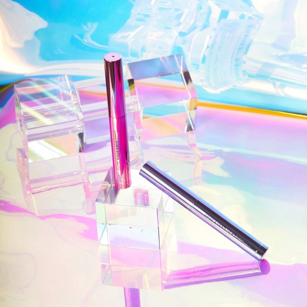 Fard à paupières Drop Eyes Stick Glitz sur blocs de verre. Emballage lilas et argenté. Fond holographique.