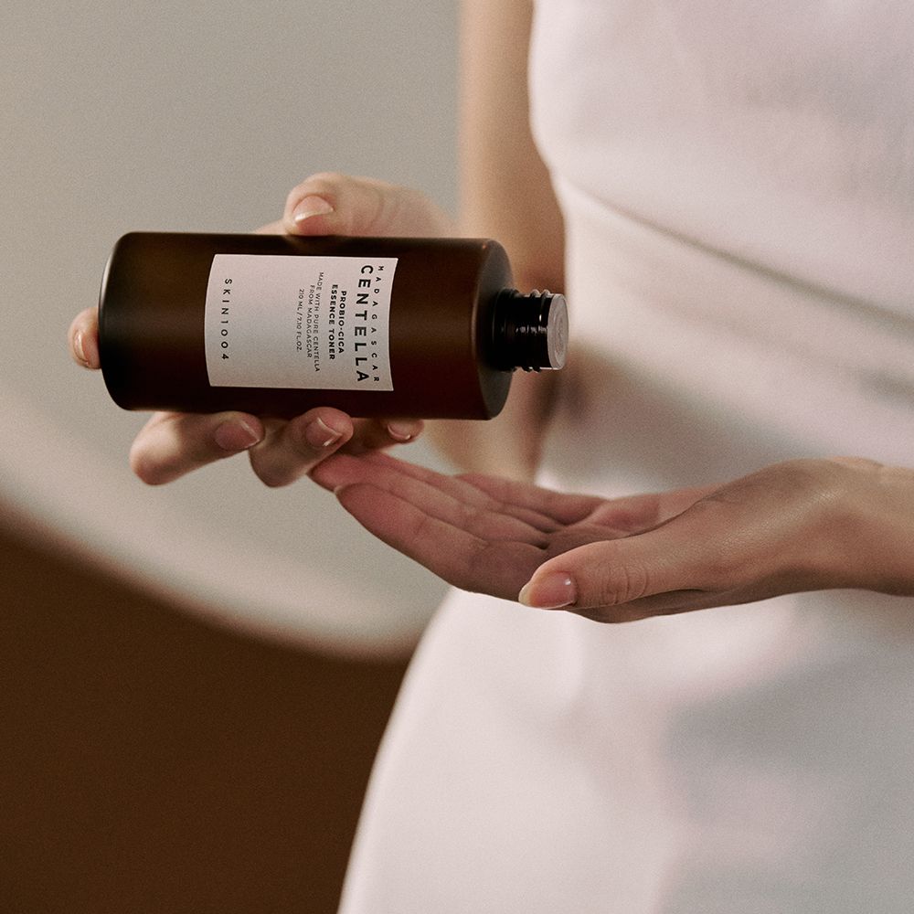 Persoon houdt bruine fles met witte dop vast. Label met tekst: CENTELLA, ESSENCE TONER, SKIN1004. Handen.