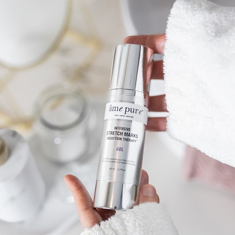 Een hand houdt een fles âme pure Intensive Stretch Mark Induction Therapy Gel vast in een badkamer.