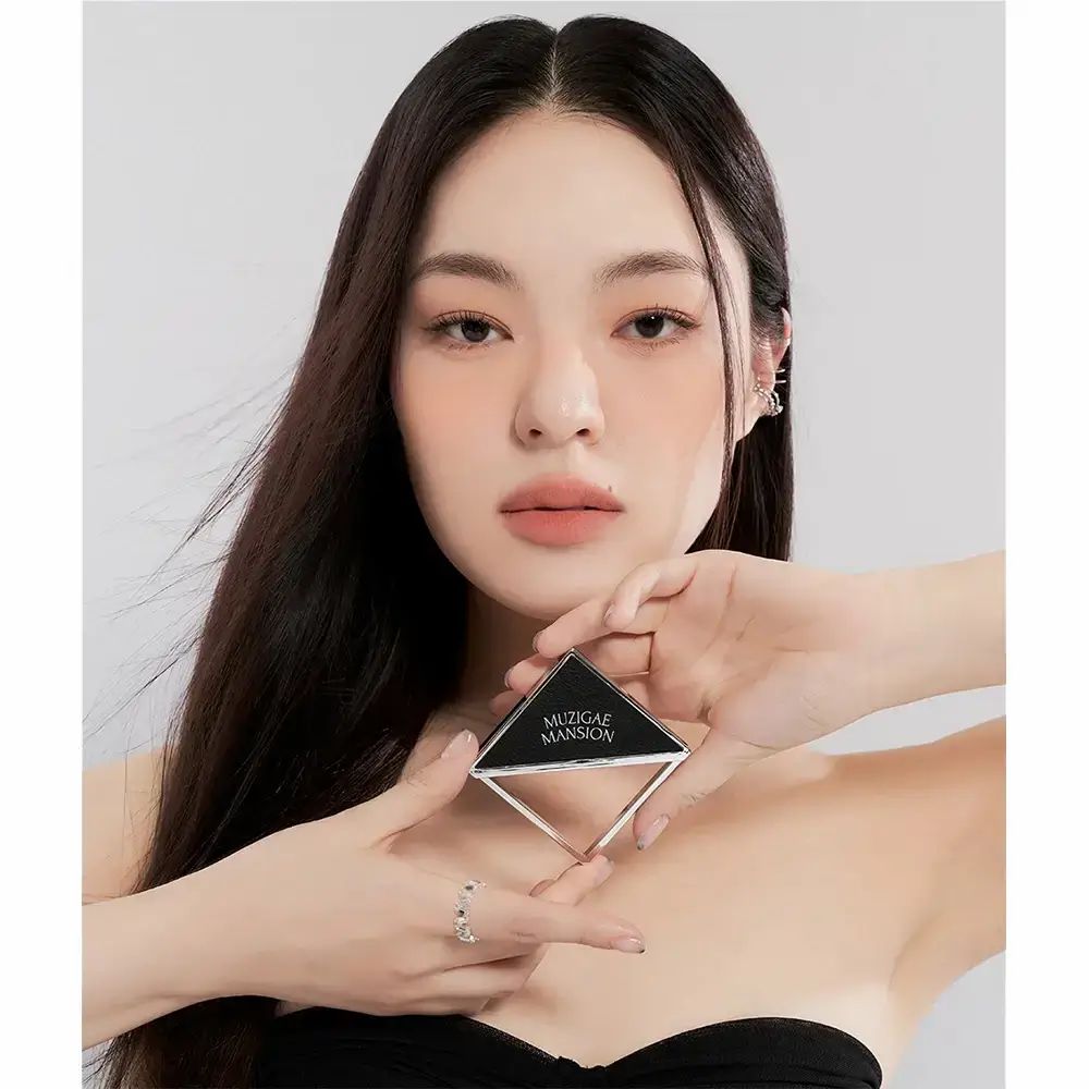 Vrouw houdt MUZIGAE MANSION blush vast. Vierkante, zilverkleurige compactverpakking. Vrouw draagt make-up.