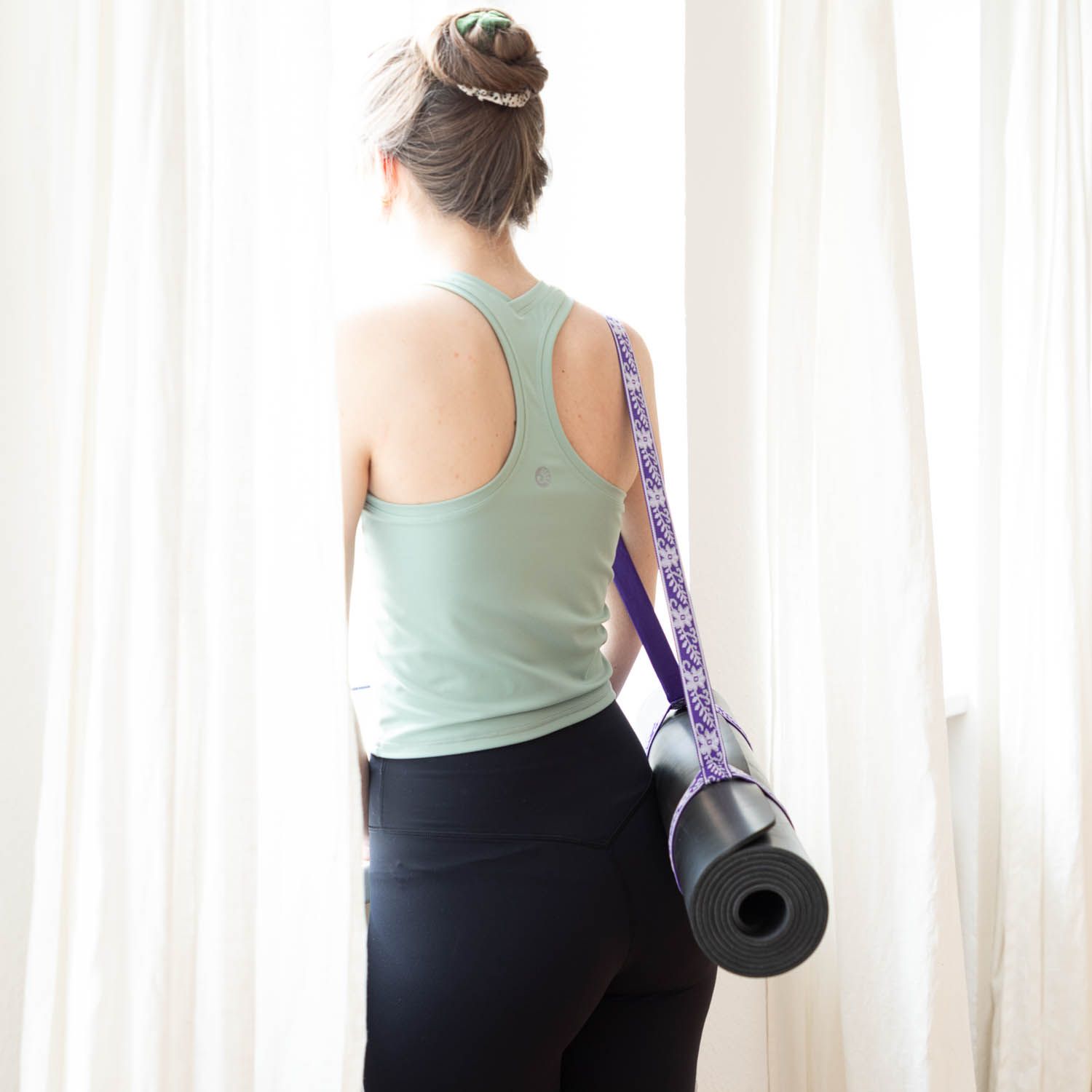 Persoon draagt yogamat met paarse draagriem. Mat opgerold. Vrouw bij raam.
