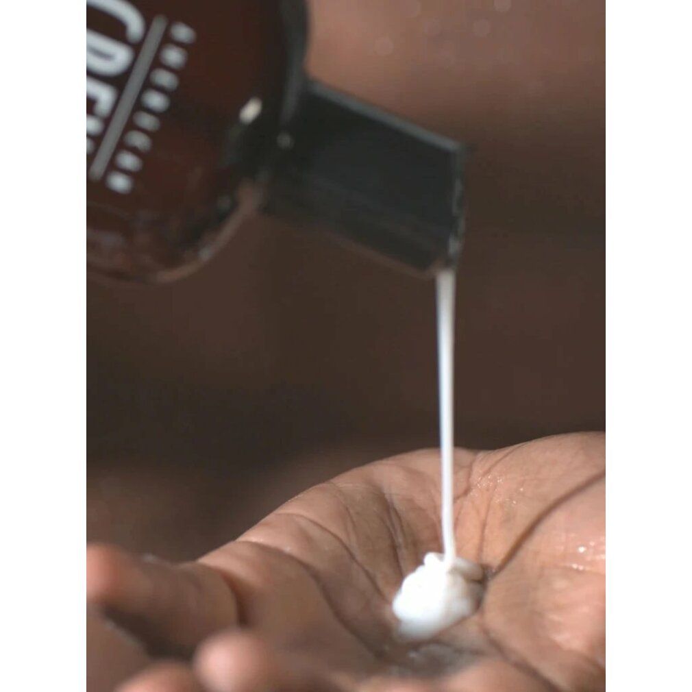 Een bruine fles met een zwarte dop, waaruit witte vloeistof in een handpalm druppelt.