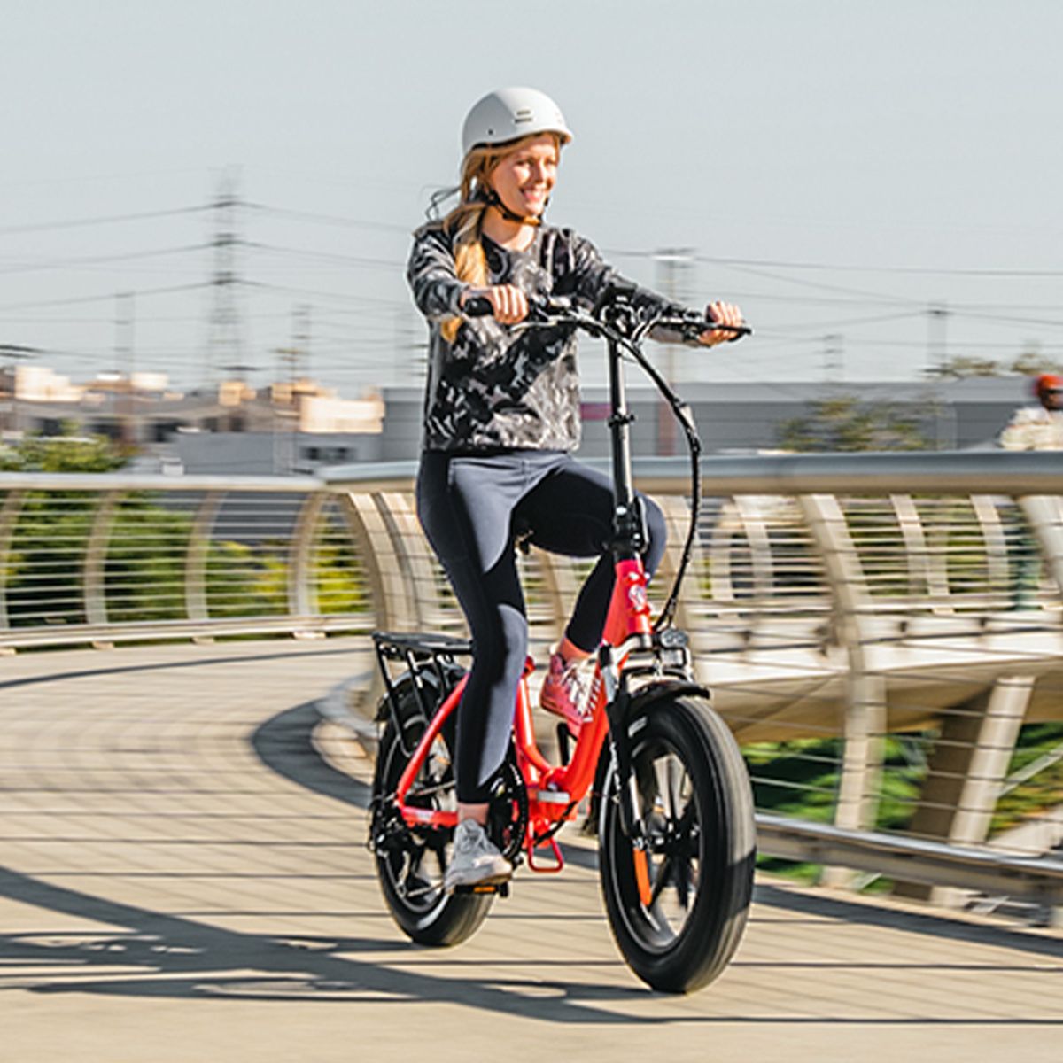 Personne faisant du vélo électrique rouge. Femme avec casque sur un pont. Arrière-plan : bâtiments et ciel. Vélo avec pneus noirs.