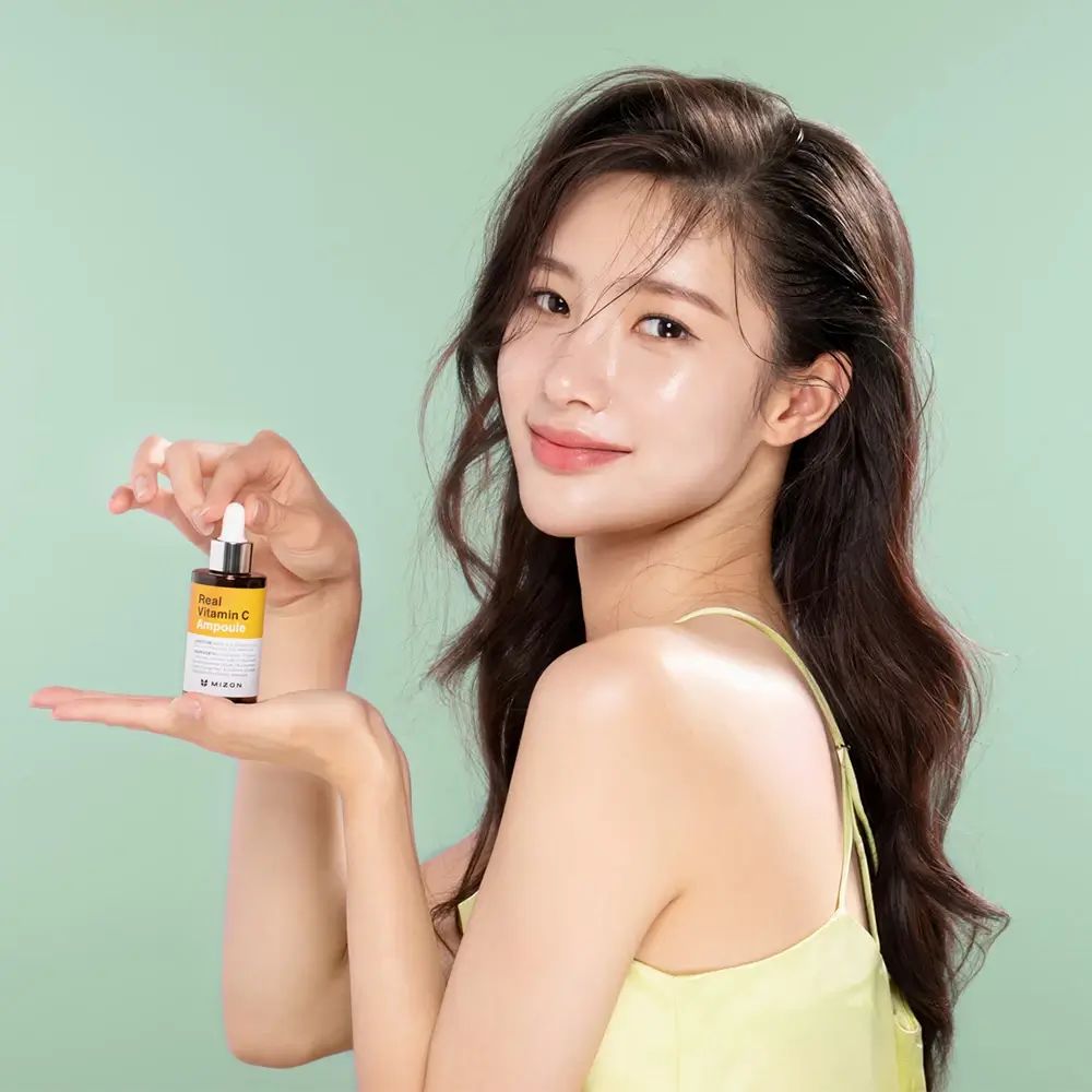 Vrouw houdt een kleine fles met geel etiket vast. Opschrift: Real Vitamin C Ampoule. Groene achtergrond.